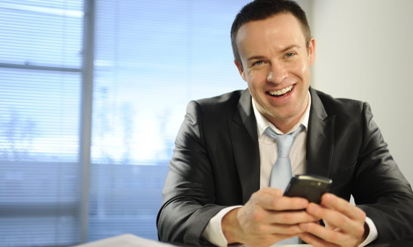 Homme souriant en costume tenant un téléphone, assis dans un bureau lumineux.